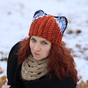 K&ouml;nnte beinhalten: Eine Frau mit langen, lockigen roten Haaren tr&auml;gt eine geh&auml;kelte M&uuml;tze mit Fuchs-Ohren. Die M&uuml;tze ist orange, wei&szlig; und schwarz. Sie tr&auml;gt einen braunen Schal und eine schwarze Jacke.