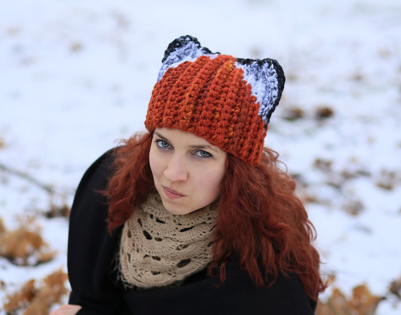 K&ouml;nnte beinhalten: Eine Frau mit langen, lockigen roten Haaren tr&auml;gt eine geh&auml;kelte M&uuml;tze mit Fuchs-Ohren. Die M&uuml;tze ist orange, wei&szlig; und schwarz. Sie tr&auml;gt einen braunen Schal und eine schwarze Jacke.