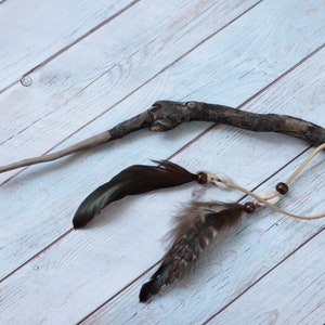 Puede incluir: Un palo de madera marrón con plumas y cuentas atadas a él. El palo está atado con un cordón de cuero marrón y tiene cuentas y conchas blancas.