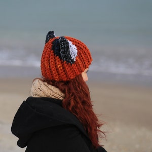 K&ouml;nnte beinhalten: Eine Frau tr&auml;gt eine rote, schwarze und wei&szlig;e H&auml;kelm&uuml;tze mit Fuchsohren. Die M&uuml;tze sitzt auf ihrem Kopf und sie schaut zur Seite. Sie steht an einem Strand mit dem Meer im Hintergrund.