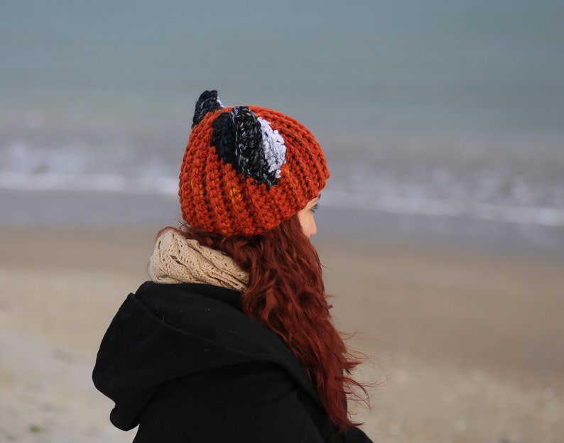 K&ouml;nnte beinhalten: Eine Frau tr&auml;gt eine rote, schwarze und wei&szlig;e H&auml;kelm&uuml;tze mit Fuchsohren. Die M&uuml;tze sitzt auf ihrem Kopf und sie schaut zur Seite. Sie steht an einem Strand mit dem Meer im Hintergrund.