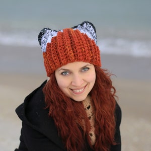 K&ouml;nnte beinhalten: Eine Frau mit langen roten Haaren tr&auml;gt eine geh&auml;kelte M&uuml;tze mit Fuchs-Ohren. Die M&uuml;tze ist orange, wei&szlig; und schwarz. Sie l&auml;chelt und schaut in die Kamera.