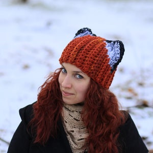 K&ouml;nnte beinhalten: Eine Frau mit langen, lockigen roten Haaren tr&auml;gt eine geh&auml;kelte M&uuml;tze mit Fuchs-Ohren. Die M&uuml;tze ist orange, wei&szlig; und schwarz. Sie tr&auml;gt eine schwarze Jacke und einen beigen Schal.