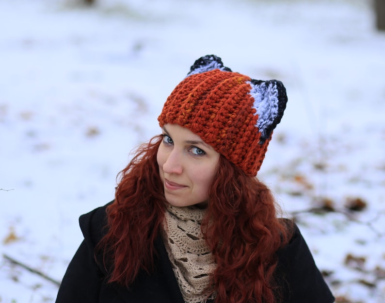 K&ouml;nnte beinhalten: Eine Frau mit langen, lockigen roten Haaren tr&auml;gt eine geh&auml;kelte M&uuml;tze mit Fuchs-Ohren. Die M&uuml;tze ist orange, wei&szlig; und schwarz. Sie tr&auml;gt eine schwarze Jacke und einen beigen Schal.