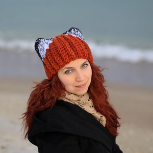 K&ouml;nnte beinhalten: Eine Frau mit langen roten Haaren tr&auml;gt eine geh&auml;kelte Fuchs-M&uuml;tze mit orangefarbenen, wei&szlig;en und schwarzen Streifen. Die M&uuml;tze hat Fuchsohren oben drauf.