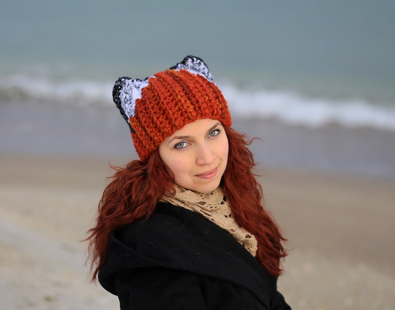 K&ouml;nnte beinhalten: Eine Frau mit langen roten Haaren tr&auml;gt eine geh&auml;kelte Fuchs-M&uuml;tze mit orangefarbenen, wei&szlig;en und schwarzen Streifen. Die M&uuml;tze hat Fuchsohren oben drauf.