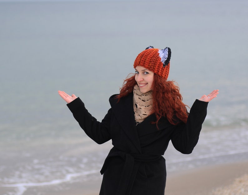 K&ouml;nnte beinhalten: Eine Frau in einem schwarzen Mantel und einer roten und wei&szlig;en Fuchs-M&uuml;tze mit Ohren, die auf einem Strand mit ausgestreckten Armen steht.