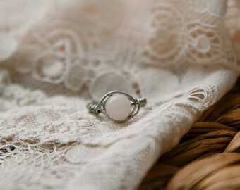 May include: A delicate silver ring with a light pink gemstone set in the center. The ring is on a white lace fabric background.