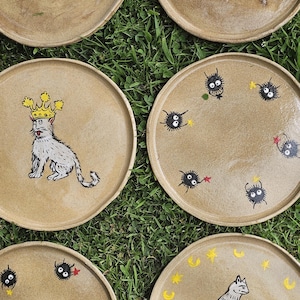 May include: A collection of six round, tan ceramic plates featuring hand-painted illustrations. Designs include a cat wearing a yellow crown, a cat holding a small creature, dinosaur skeletons, and several small, fuzzy black soot sprites with stars.