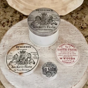 May include: A collection of four vintage-style containers including a white jar with a metal lid labeled Burgess's Genuine Anchovy Paste, a matching lid, a Woods Cherry Tooth Paste lid, and a small round container labeled Lip Salve.