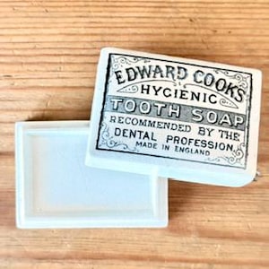 May include: A rectangular white ceramic container with a lid featuring black printed text that reads EDWARD COOKS HYGIENIC TOOTH SOAP RECOMMENDED BY THE DENTAL PROFESSION MADE IN ENGLAND, surrounded by decorative scrollwork borders.