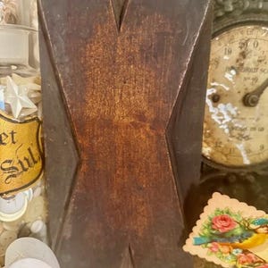 May include: A dark brown, wooden block with a carved 'X' design. The block has a rustic, aged appearance with visible wood grain. The background includes a vintage clock, a small jar with stars, and a 'Be Mine' heart.