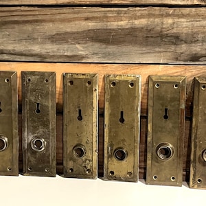 May include: Six antique brass door plates with keyholes and circular knob openings. The rectangular plates show signs of age and wear, with a warm, golden hue. The plates are arranged horizontally against a wooden background.