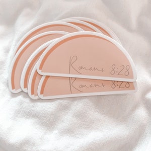 May include: A stack of peach-colored stickers with a rounded top and a white outline. The stickers have the text "Romans 8:28" written in a cursive font.