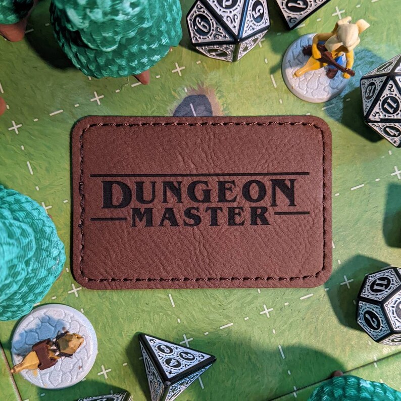 K&ouml;nnte beinhalten: Eine braune Dungeon Master Spielmatte aus Kunstleder mit gen&auml;hten Kanten. Die Matte tr&auml;gt den Schriftzug "Dungeon Master" in Schwarz.