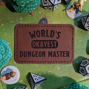 K&ouml;nnte beinhalten: Ein braunes Leder-Patch mit dem Text "World's Okayest Dungeon Master" in schwarzen Buchstaben.