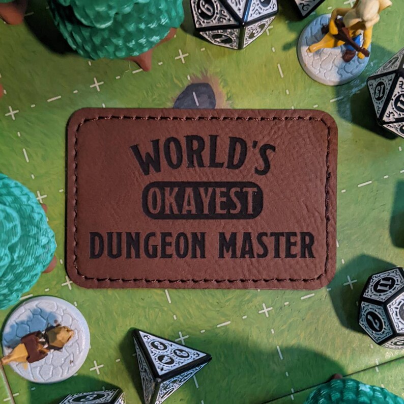 K&ouml;nnte beinhalten: Ein braunes Leder-Patch mit dem Text "World's Okayest Dungeon Master" in schwarzen Buchstaben.
