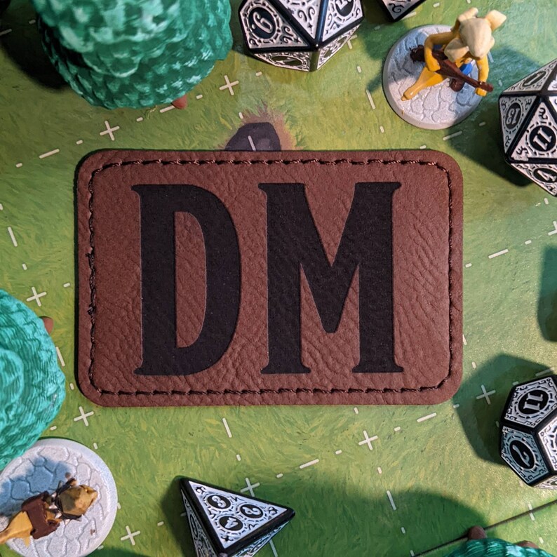 K&ouml;nnte beinhalten: Braunes Leder-Patch mit den Buchstaben DM in Schwarz, umgeben von wei&szlig;en N&auml;hten. Das Patch befindet sich auf einer gr&uuml;n-wei&szlig;en Oberfl&auml;che mit W&uuml;rfeln und einer Miniaturfigur.