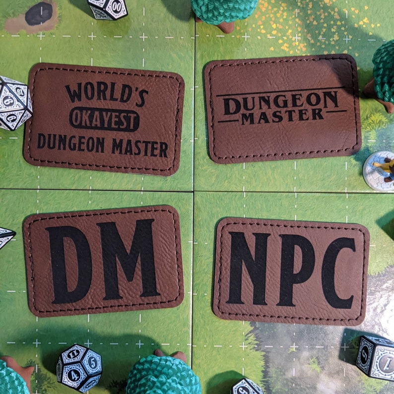 K&ouml;nnte beinhalten: Vier braune Leder-Patches mit schwarzem Text. Die Patches lauten "World's Okayest Dungeon Master", "Dungeon Master", "DM" und "NPC".