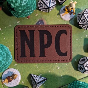 K&ouml;nnte beinhalten: Ein braunes Leder-Patch mit dem Wort "NPC" in schwarzen Buchstaben gedruckt. Das Patch ist von W&uuml;rfeln und anderen Spielsteinen auf einer gr&uuml;nen Filzoberfl&auml;che umgeben.