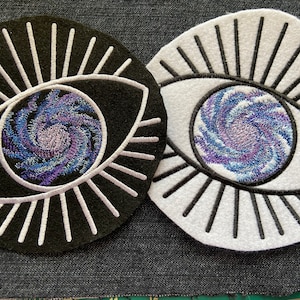 May include: Two embroidered patches, one black and one white, each featuring a stylized eye with a swirling galaxy design in shades of blue, purple, and white.