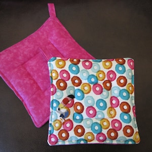 May include: Two pink and white pot holders. One is solid pink with a quilted design. The other is white with a donut pattern and a toucan illustration.
