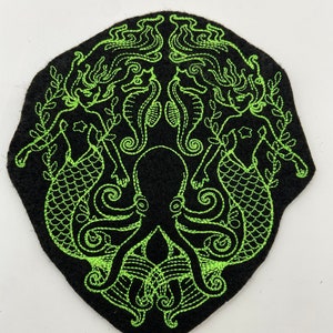 May include: A black felt patch with an embroidered design of two mermaids flanking an octopus. The mermaids have flowing hair and tails, and the octopus has eight tentacles. The design is outlined in green thread.