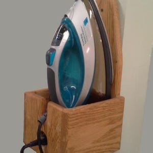 May include: A white and teal iron with a black soleplate rests in a wooden holder. The holder is mounted on a wall and has a slot for the iron's power cord. The iron is designed for home use.