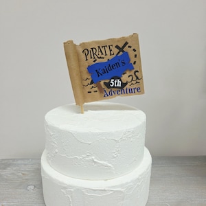May include: A two-tiered white cake with a pirate-themed cake topper. The topper is a scroll with the words "PIRATE" and "Kaiden's 5th Adventure" in blue and black. The cake has a textured frosting.