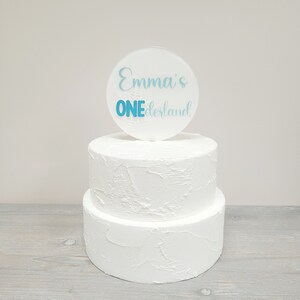 May include: A two-tiered white cake with a textured finish, crowned with a clear, circular acrylic cake topper. The topper displays the text "Emma's ONEderland" in light blue script and block letters, with snowflake details.