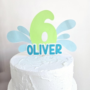 Puede incluir: Un adorno para pastel de color verde con el número seis, salpicaduras de agua azules y el nombre "OLIVER" en letras azules.