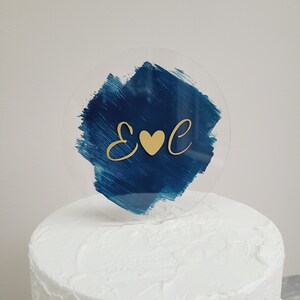 Puede incluir: Un adorno para pastel circular transparente con un diseño de pincelada azul marino. Las letras doradas "E" y "C" flanquean un corazón dorado. El adorno se coloca encima de un pastel glaseado blanco, creando una estética festiva.