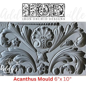 May include: A gray silicone craft mould by Iron Orchid Designs featuring intricate acanthus leaf scrolls, a central shell motif, small flowers, and a beaded border. The rectangular mould measures 6 inches by 10 inches and is labeled Acanthus Mould.