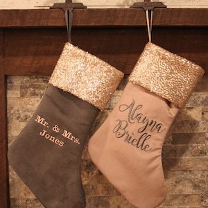 May include: Two personalized Christmas stockings with gold sequin trim. The stocking on the left is gray with the text "Mr. & Mrs. Jones" embroidered on it. The stocking on the right is pink with the text "Alayna Brielle" embroidered on it.