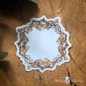 May include: A white sticker with a brown design of four hares hopping in a circle around a vine. The hares are facing inward and their tails are curled. The sticker is on a wooden surface.