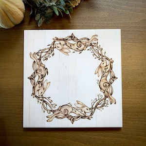 May include: A white wooden plaque with a design of four brown rabbits in a circle of vines. The rabbits are facing inwards towards the center of the circle.