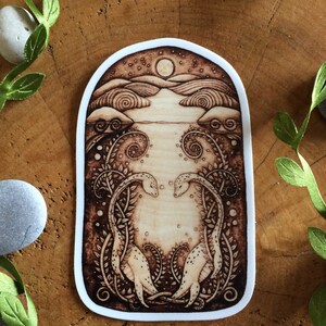 Puede incluir: Una pegatina quemada en madera con un dise&ntilde;o caprichoso de dos criaturas estilizadas con cuellos largos, que se miran en un paisaje iluminado por la luna. El fondo presenta un cielo estrellado, colinas y una masa de agua.
