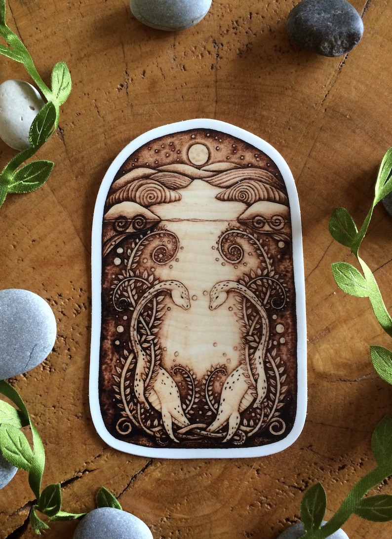Puede incluir: Una pegatina quemada en madera con un dise&ntilde;o caprichoso de dos criaturas estilizadas con cuellos largos, que se miran en un paisaje iluminado por la luna. El fondo presenta un cielo estrellado, colinas y una masa de agua.
