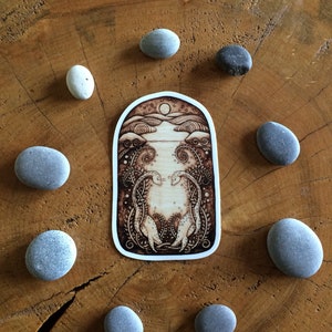 Puede incluir: Una pegatina quemada en madera con un dise&ntilde;o caprichoso que presenta dos criaturas estilizadas con cuellos y colas largos, rodeadas de enredaderas en espiral y una luna en el cielo. La pegatina est&aacute; sobre una superficie de madera con piedras grises alrededor.