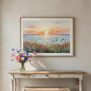 Könnte beinhalten: Ein gerahmtes Aquarellgemälde eines Sonnenuntergangs über dem Meer, mit bunten Wildblumen im Vordergrund. Das Gemälde hängt über einer weißen, rustikalen Holzkonsolentisch mit einer geflochtenen Bank und einer Blumenvase.