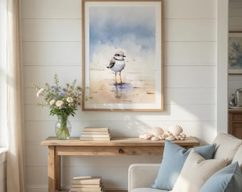 May include: A framed watercolour print of a small shorebird on a beach. The artwork uses soft blues and tans, with the bird's reflection in the wet sand. The print hangs above a wooden console table with books, flowers, and seashells, creating a coastal theme.