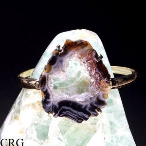 May include: A gold bangle bracelet with a large, natural agate geode. The geode is brown, black, and white with a hollow center. The bracelet is displayed on a green rock.