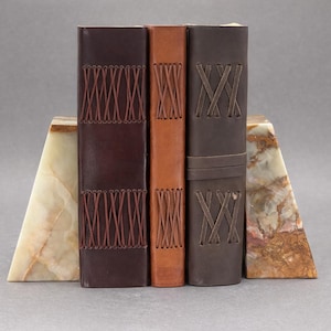 May include: A set of three leather-bound journals in shades of brown, with decorative stitching. The journals are held between two triangular onyx bookends with cream and brown marbling.