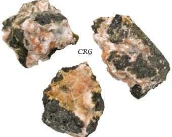 Puede incluir: Tres rocas rugosas, de forma irregular, de color verde oscuro y naranja. Las rocas tienen una superficie rugosa y texturizada y están cubiertas de pequeños cristales blancos.