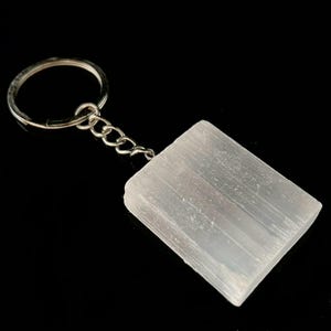 Puede incluir: Un llavero de cristal de selenita blanco con un anillo y una cadena de metal plateado. El cristal es de forma rectangular y tiene una superficie lisa y pulida.