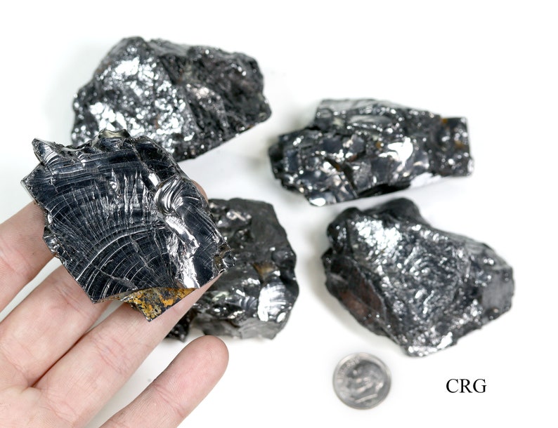 May include: Five pieces of raw, black, metallic, rough, and jagged,  cobalt ore. The ore is displayed on a white surface with a US dime for scale. The text "CRG" is printed in black on the white surface.