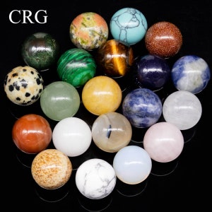 May include: A collection of 17 smooth, polished gemstone spheres in various colors and patterns, including green, blue, red, brown, white, and black. The spheres are arranged in a circular pattern on a black surface.