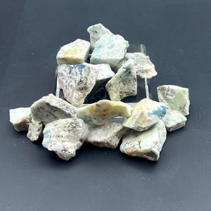 May include: A collection of rough-cut, natural stones in shades of white, gray, and pale green, with accents of blue and yellow. The stones are irregularly shaped and appear to be raw minerals, possibly for crystal healing or decorative purposes.