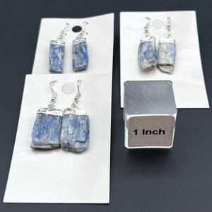 May include: Four pairs of blue kyanite earrings with silver-colored metal accents. The earrings are displayed on white cards. A silver-colored cube with the text "1 Inch" is also in the image.