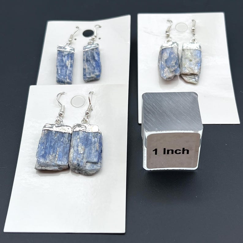 May include: Four pairs of blue kyanite earrings with silver-colored metal accents. The earrings are displayed on white cards. A silver-colored cube with the text "1 Inch" is also in the image.